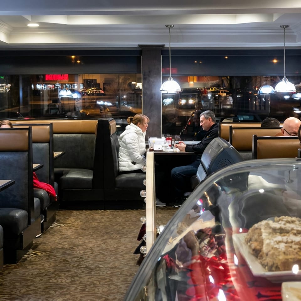 Cozy diner interior with illuminated booths, seated patrons, pendant lights, and a glass display case of baked goods.