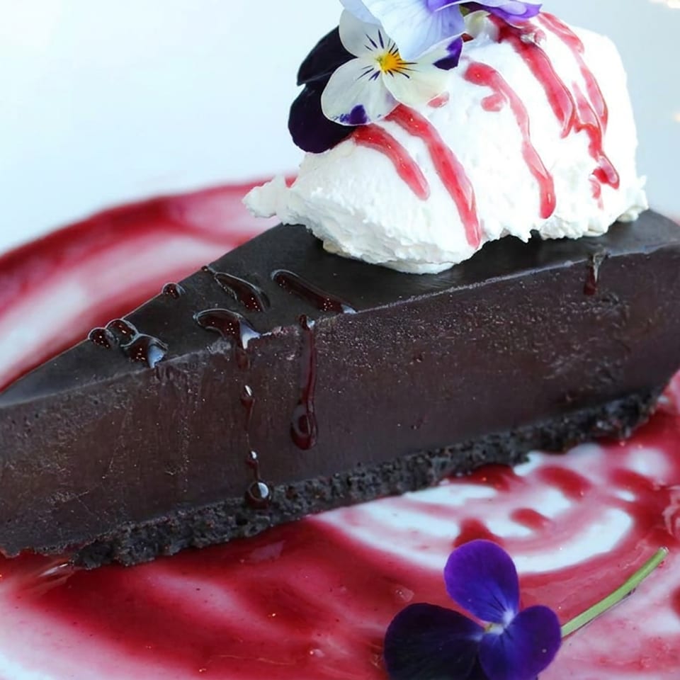Rich chocolate cake topped with whipped cream, raspberry sauce, and edible purple flowers on a decorative plate.