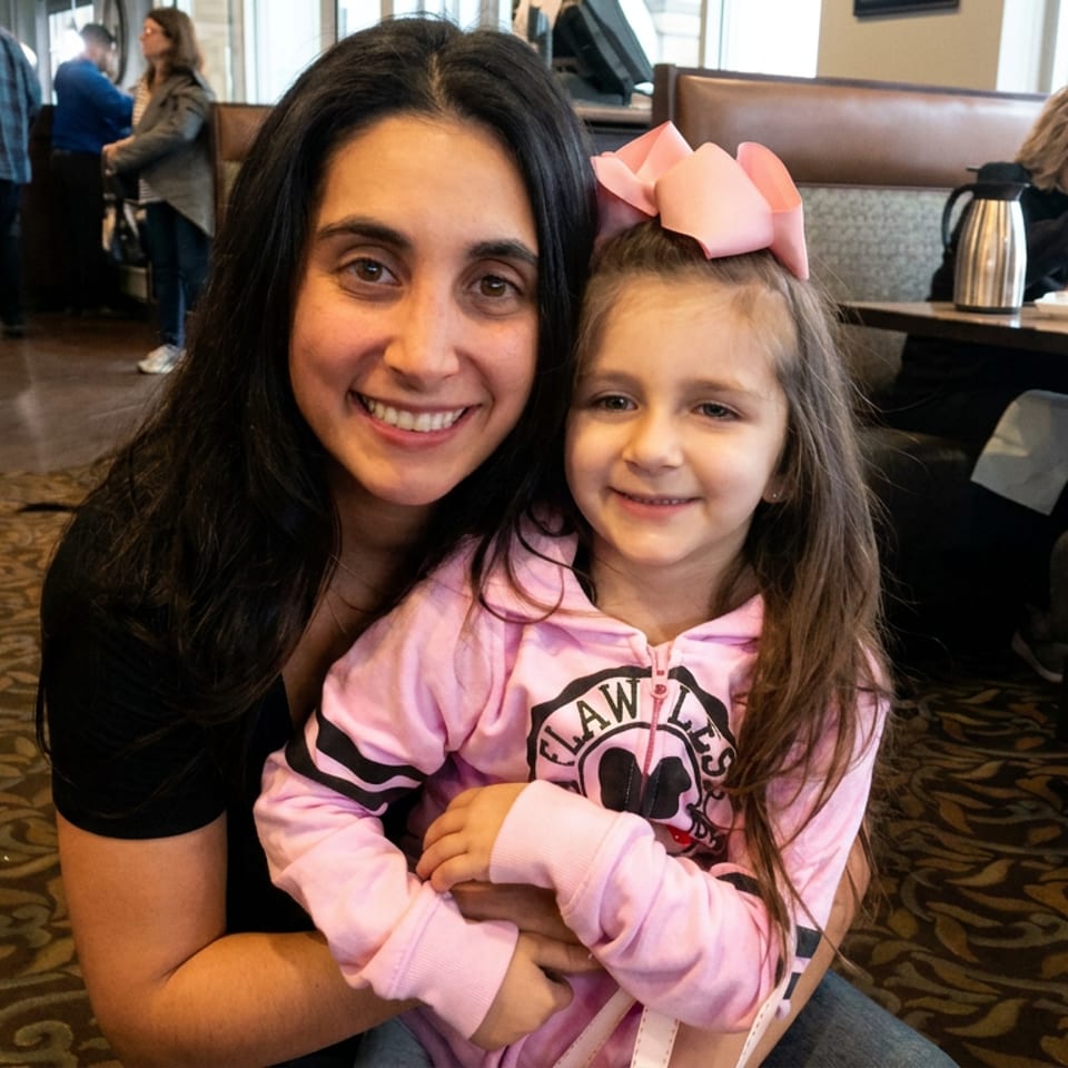 Smiling woman hugs a young girl wearing a pink bow inside a warmly lit restaurant dining area.
