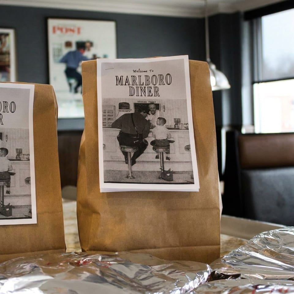 Two Marlboro Diner takeout bags and foil-wrapped meals sit on a counter inside a warmly lit diner.